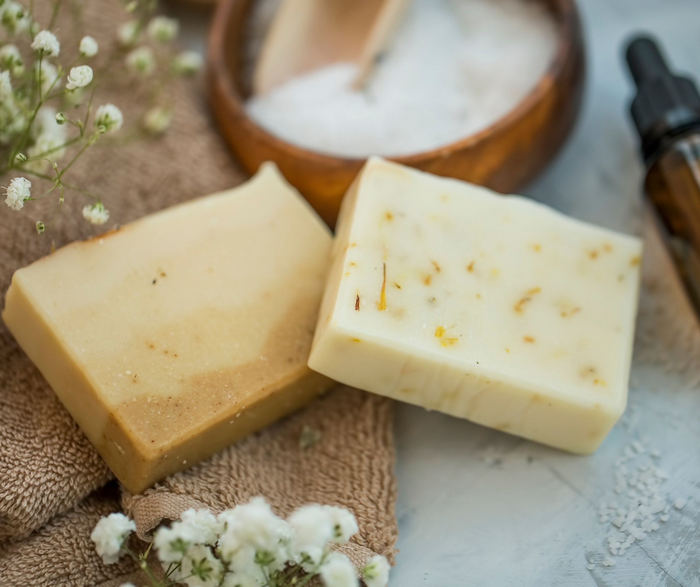Two soap bars on a textured surface with a wooden bowl and dropper in the background.